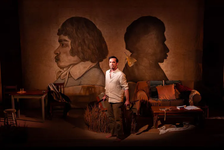 A performer stands in a dimly-lit living room set beneath two large profile portraits during a theatre performance.