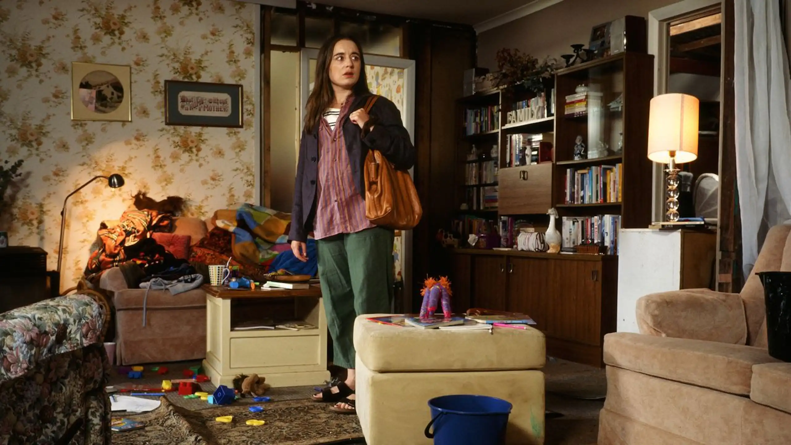 A movie scene in which a young woman stand inside a messy lounge room with a brown purse.