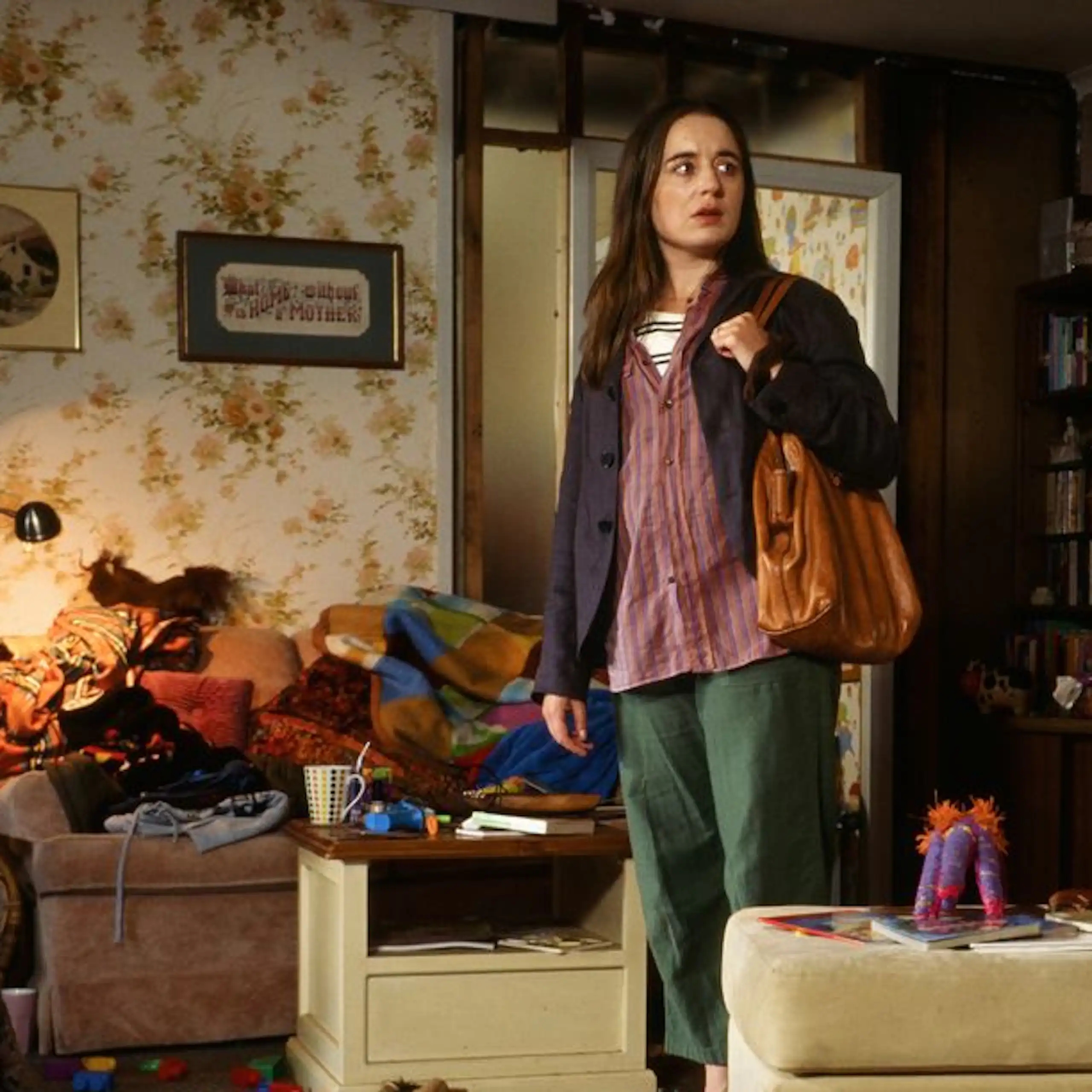 A movie scene in which a young woman stand inside a messy lounge room with a brown purse.