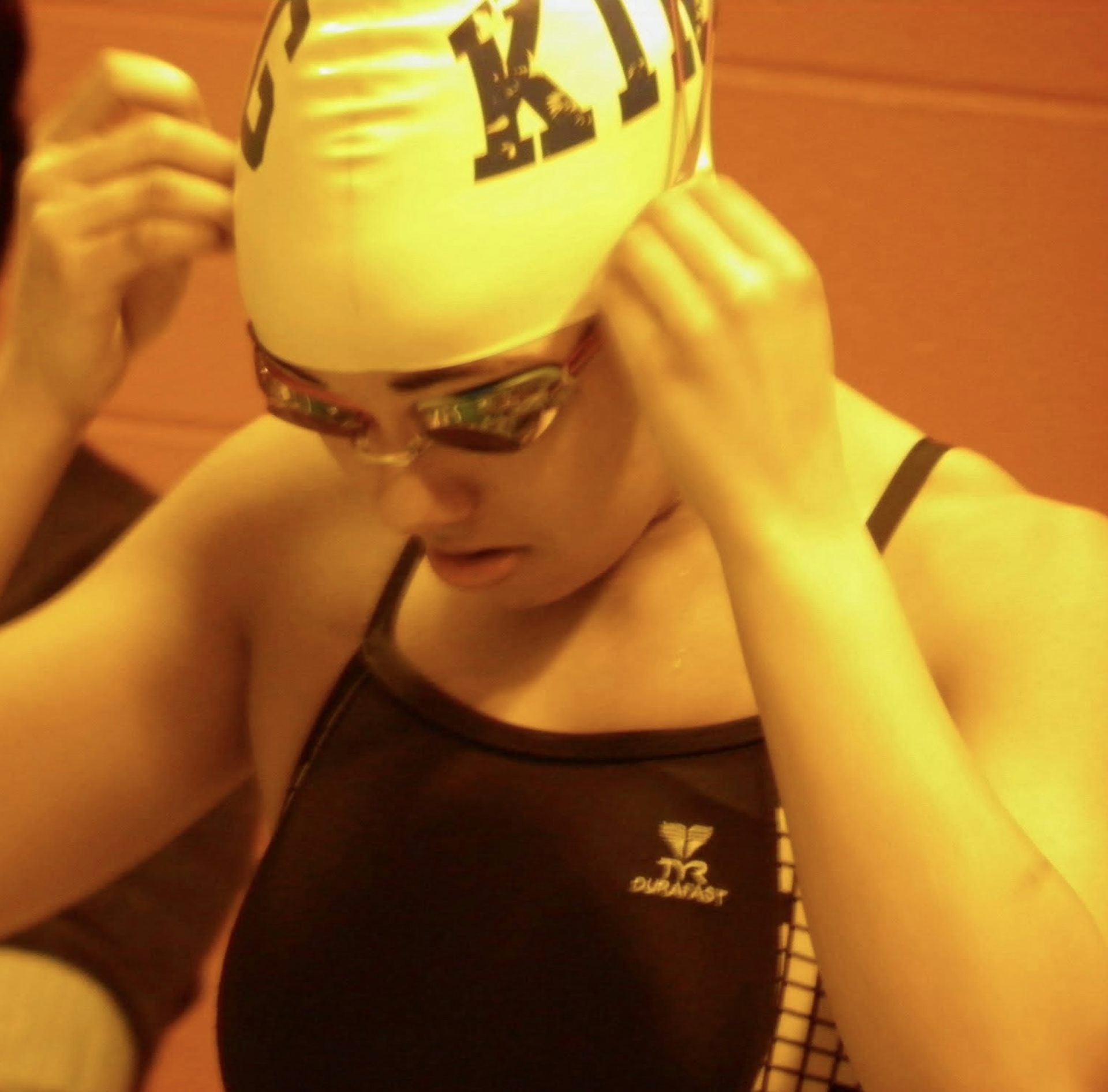 Orange-toned photo of teen in swimsuit and swimcap adjusting goggles