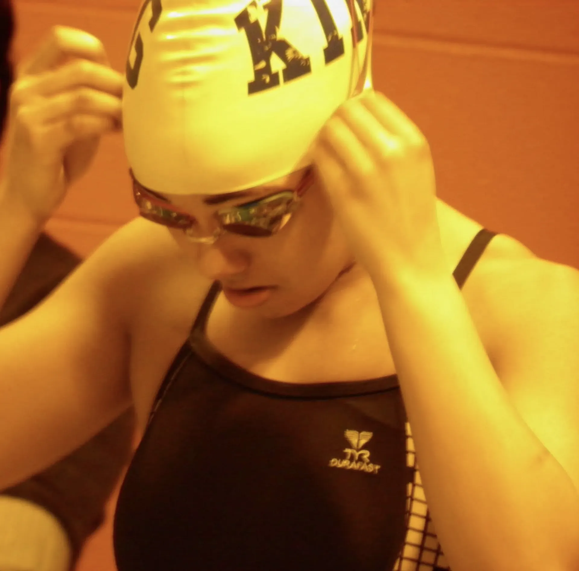 Orange-toned photo of teen in swimsuit and swimcap adjusting goggles