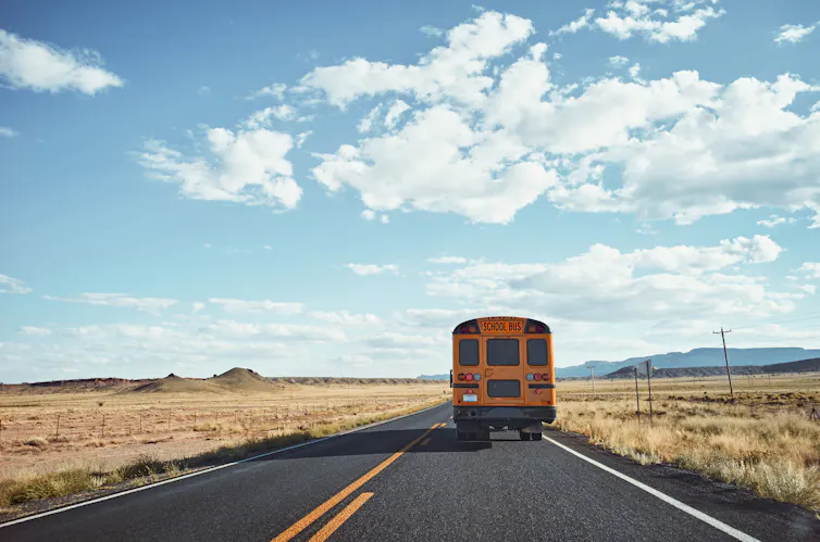 A yellow school bus is seen on an empty highway in a desert with a blue sky and white clouds.