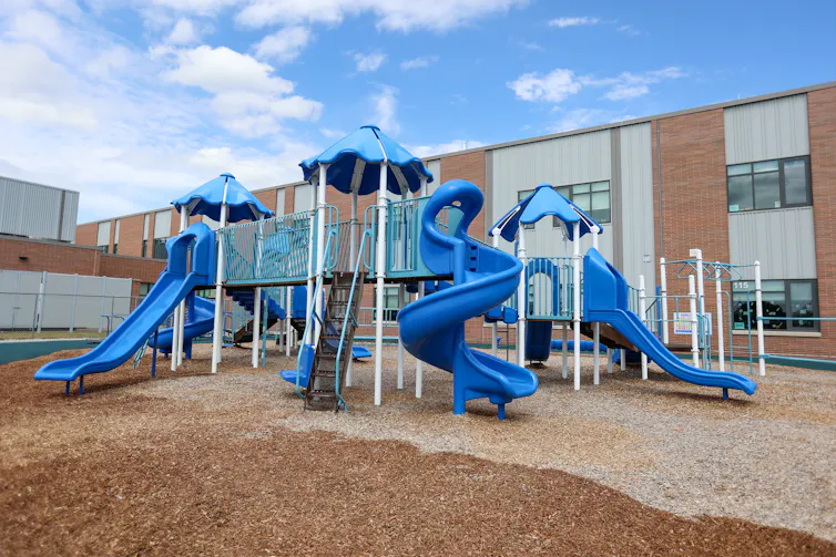 A modern-looking blue playground is empty outside of a large brown and white building.