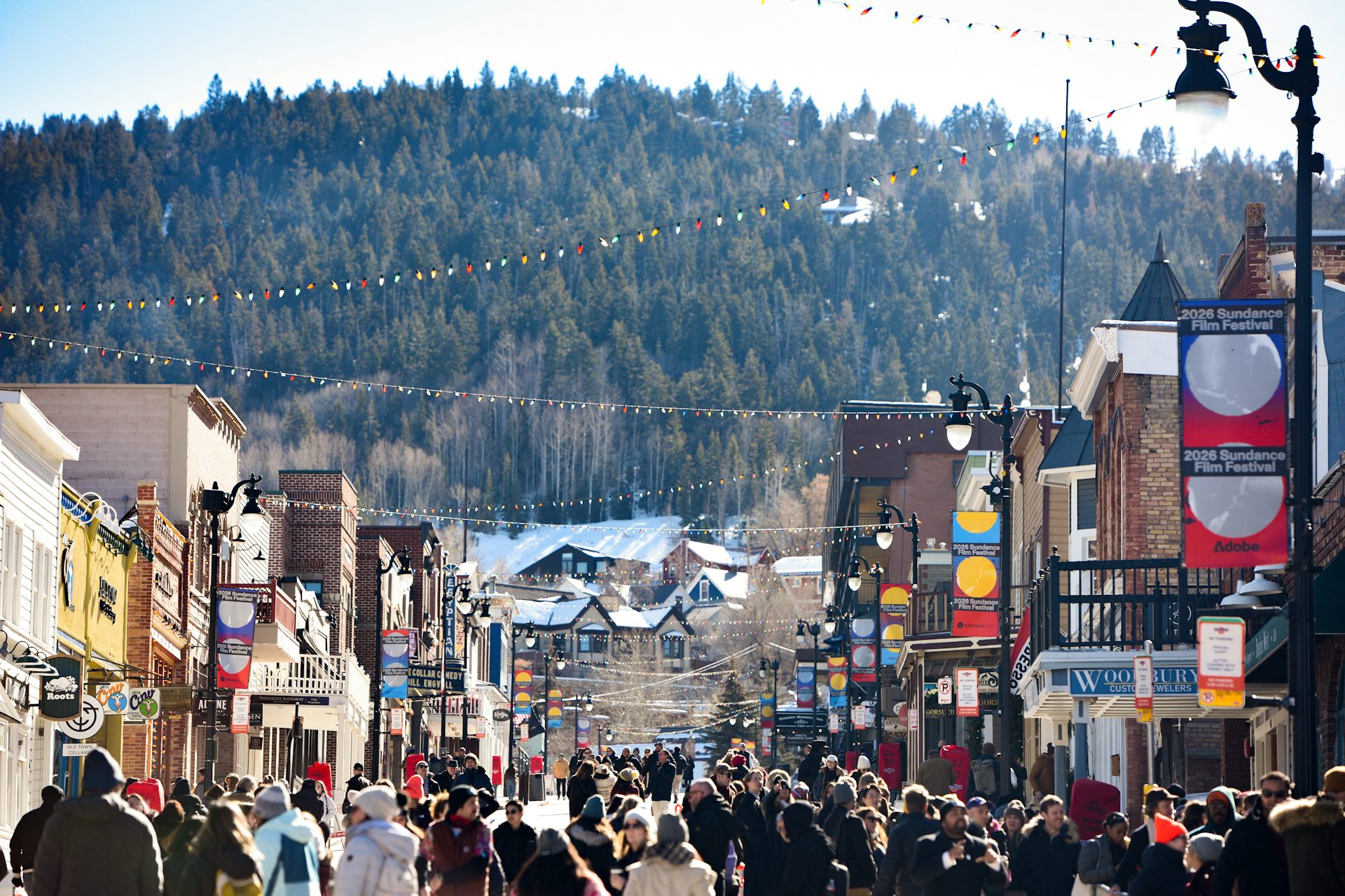 A crowd of people walk on a street with two-story buildings on either side and lights strung overhead. A mountain filled with trees looms over them in the background.