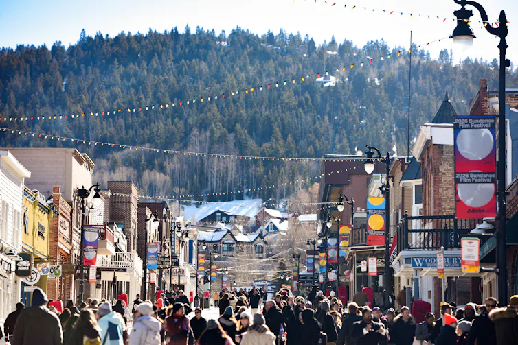 A crowd of people walk on a street with two-story buildings on either side and lights strung overhead. A mountain filled with trees looms over them in the background.