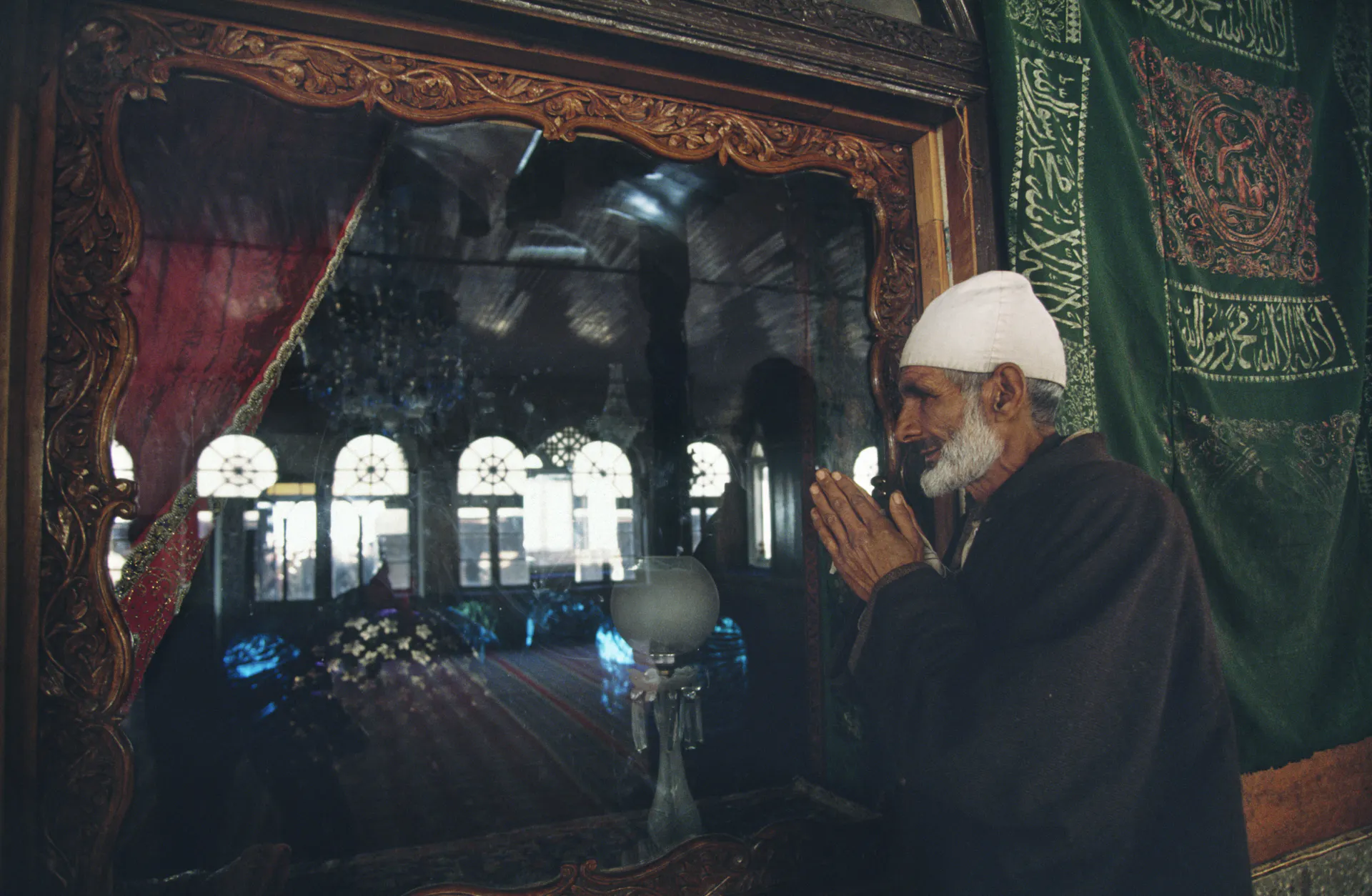 A man stands outside an ornate doorway with curtains pinned to either side, his hands folded in prayer.