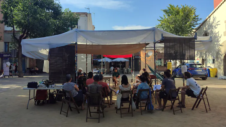 Arquitectura temporal d'ombra en una plaça oberta, feta amb bastides i teles, sota la qual té lloc una conversa pública amb gent asseguda en cadires.