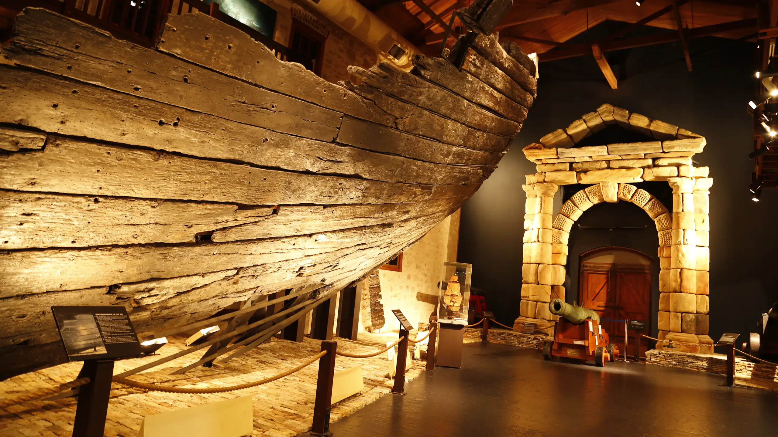 A weathered timber ship hill in a museum, alongside a stone archway.