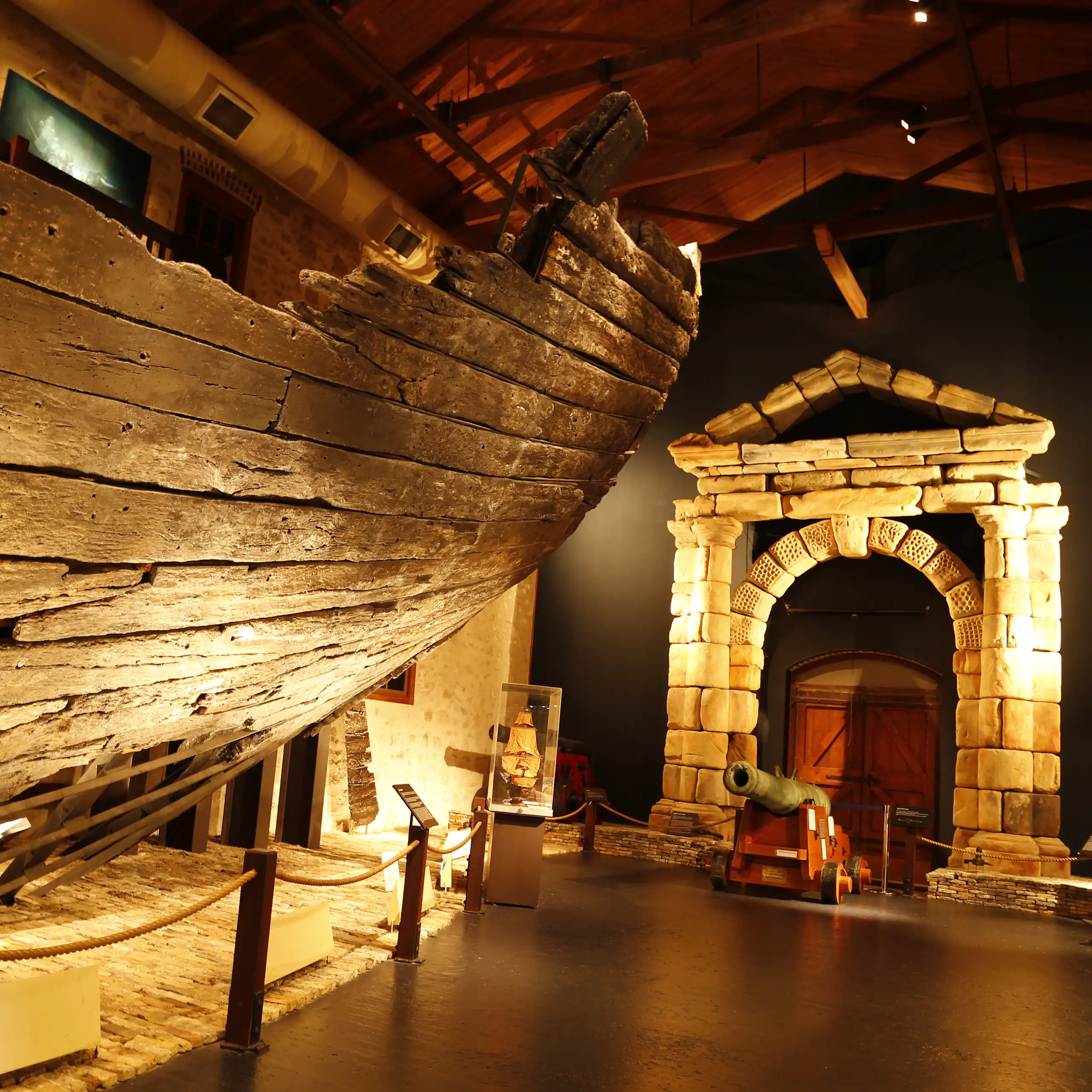 A weathered timber ship hill in a museum, alongside a stone archway.