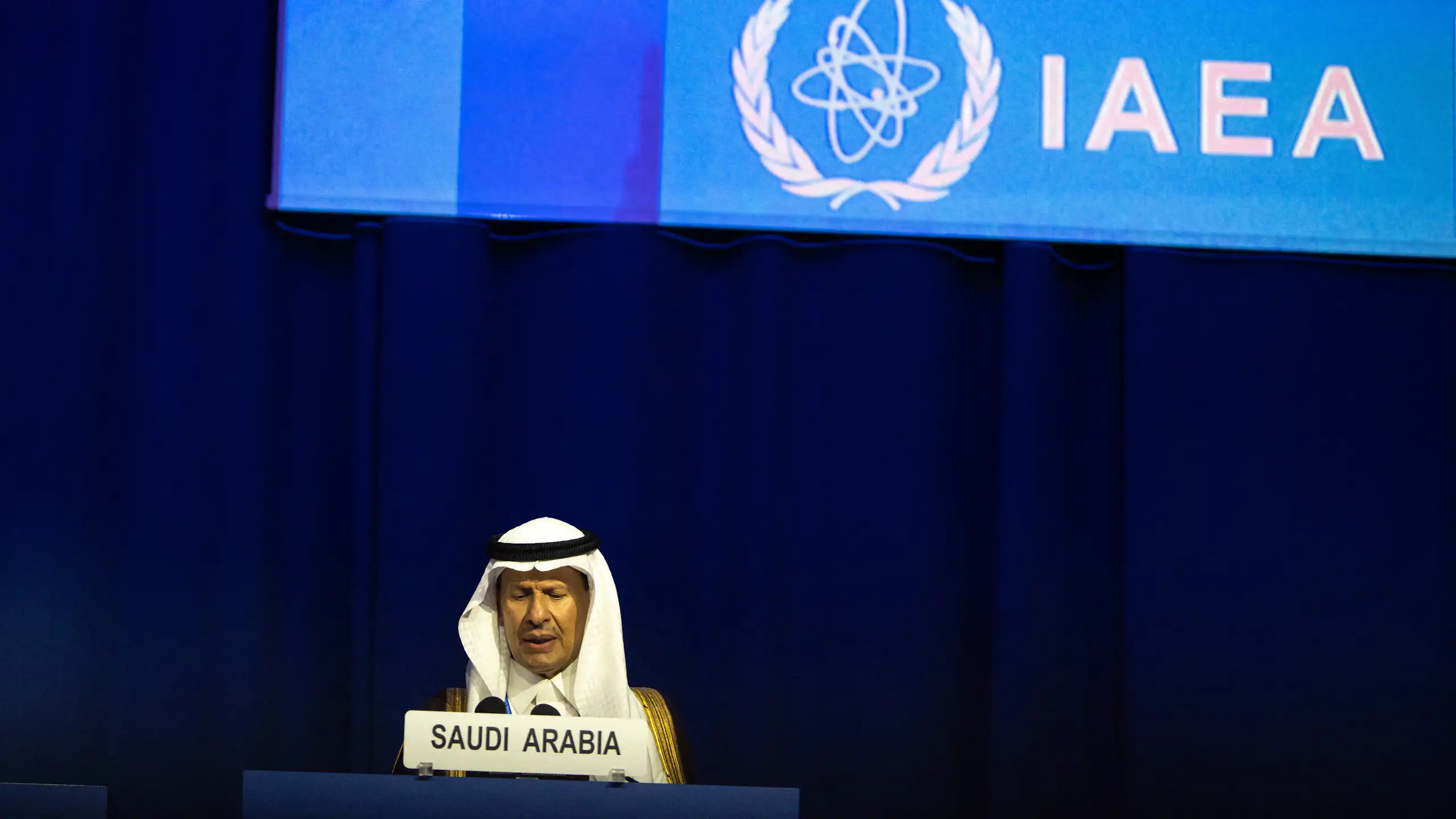 A man sits at a lectern in front of a large screen with IAEA written on it.