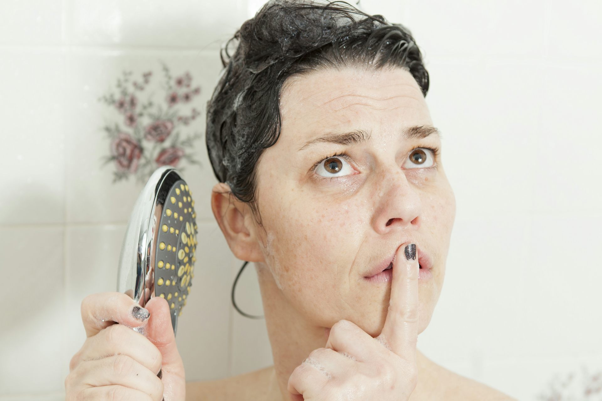 A woman holds a shower head in one hand while staring at the ceiling in deep thought.