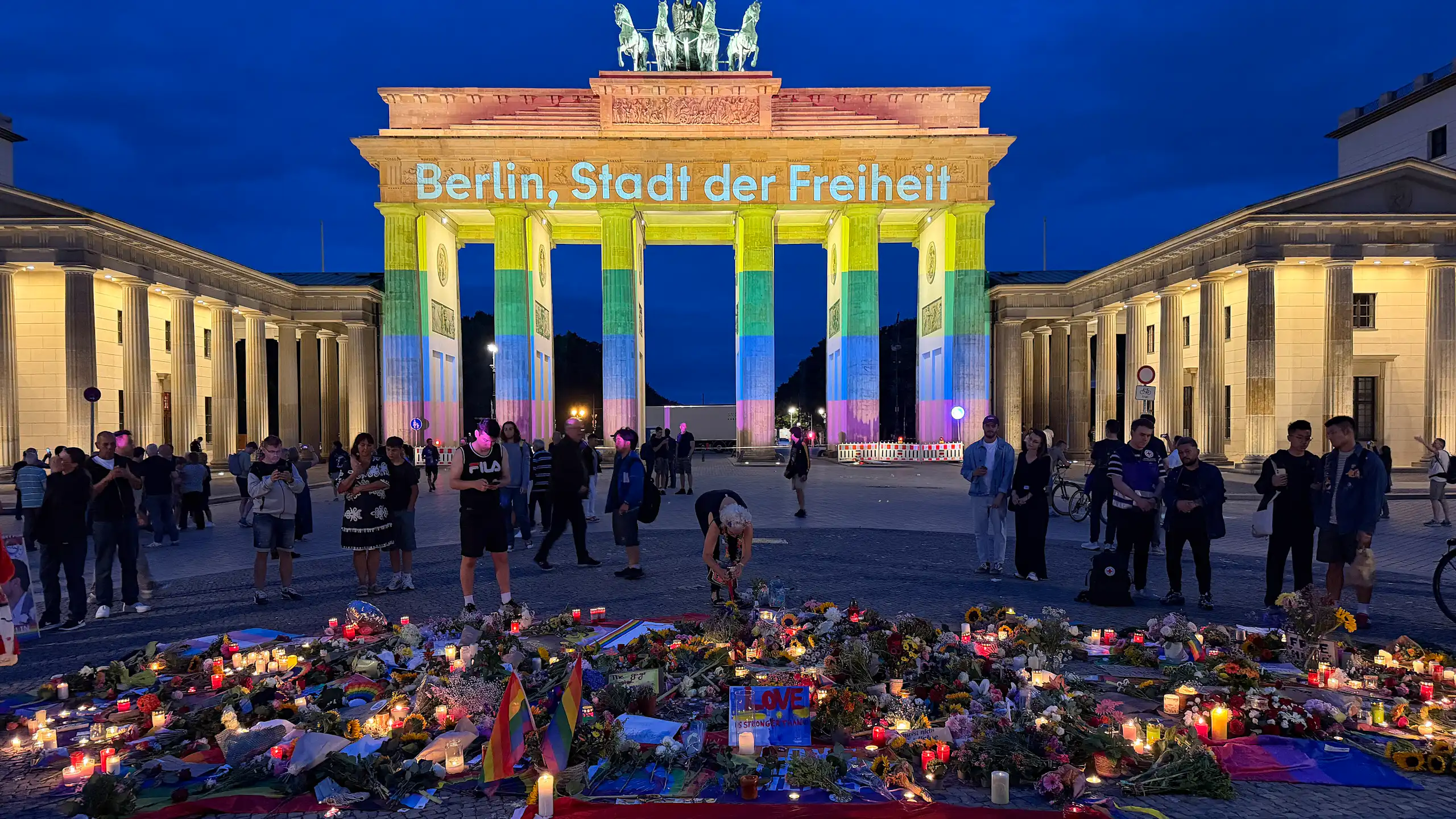 Berlin's Brandenburg Gate is lit up in the colours of the rainbow flag, with flowers, candles and rainbow flags on the ground in front of it.