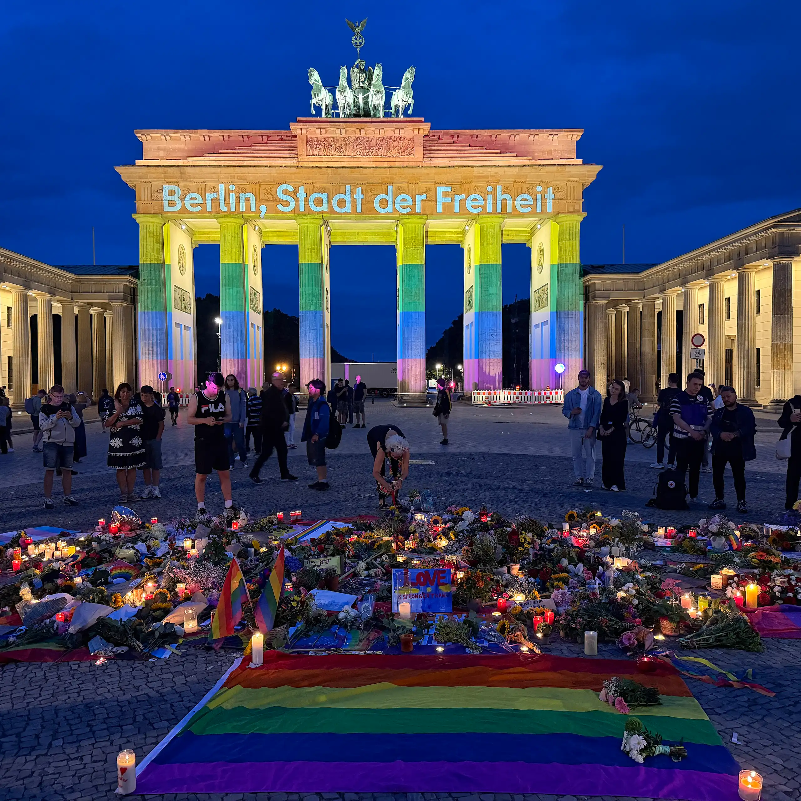 Berlin's Brandenburg Gate is lit up in the colours of the rainbow flag, with flowers, candles and rainbow flags on the ground in front of it.
