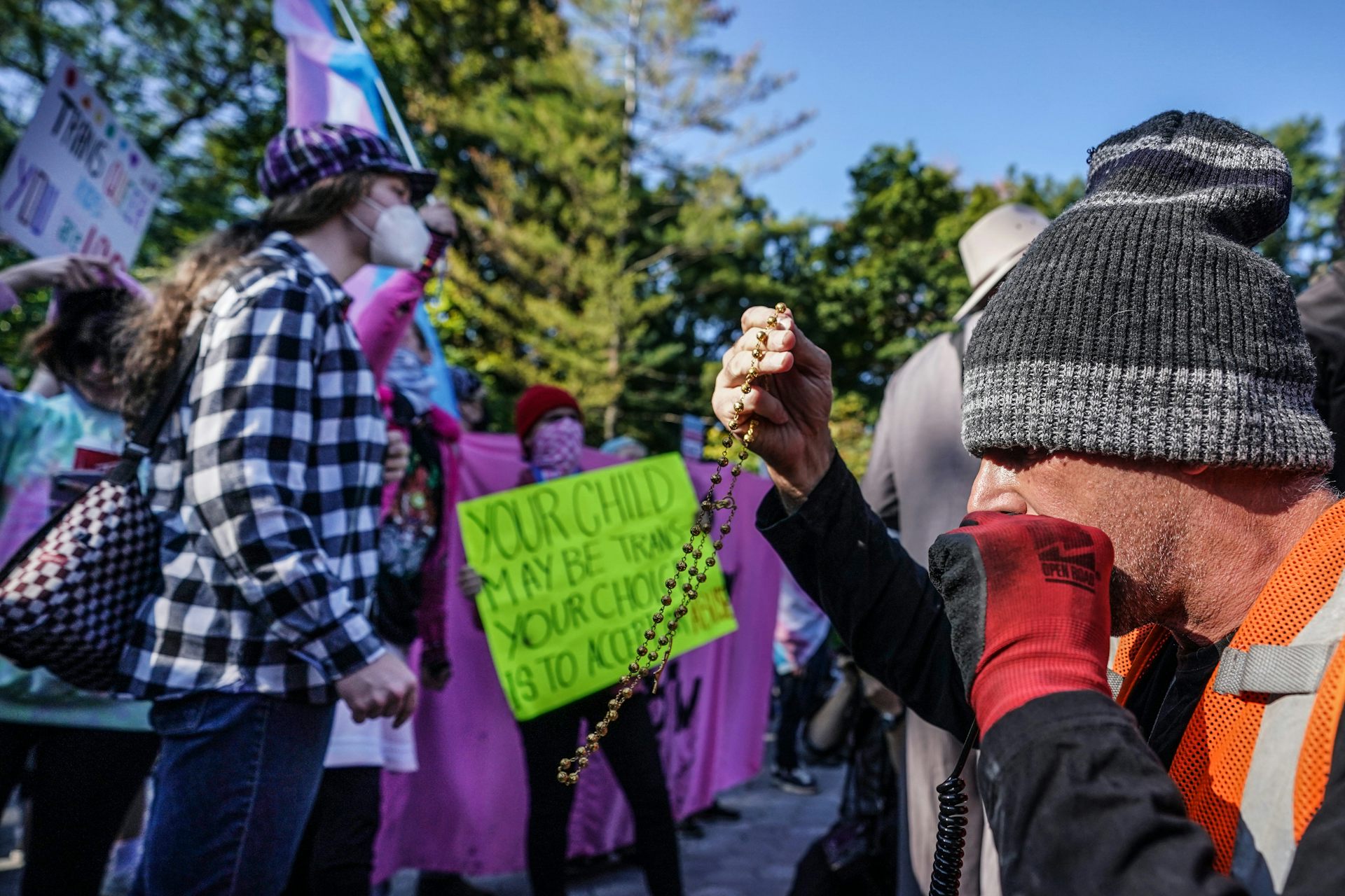 A protester holds a rosary and recites prayers next to a group of people counter-protesting and holding signage and flags.