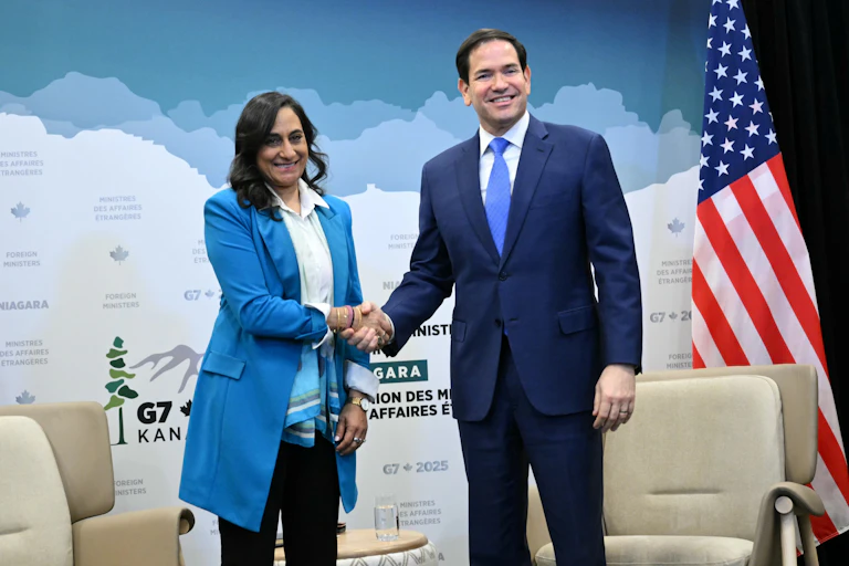 A woman with dark hair in a blue jacket shakes hands with a dark-haired man in a suit.