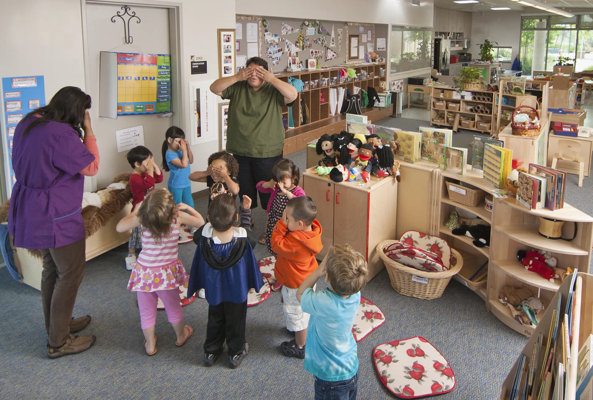 Young children cover their eyes, alongside adults who do the same in a colorful classroom.