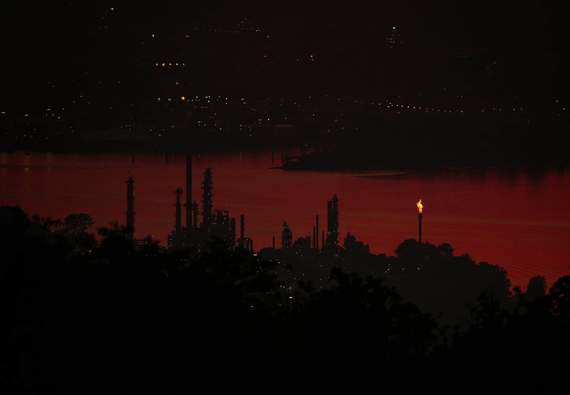 Eine Ölraffinerie mit einer Fackel in der Nähe eines Flusses mit einem Stadtbild im Hintergrund bei Sonnenuntergang.