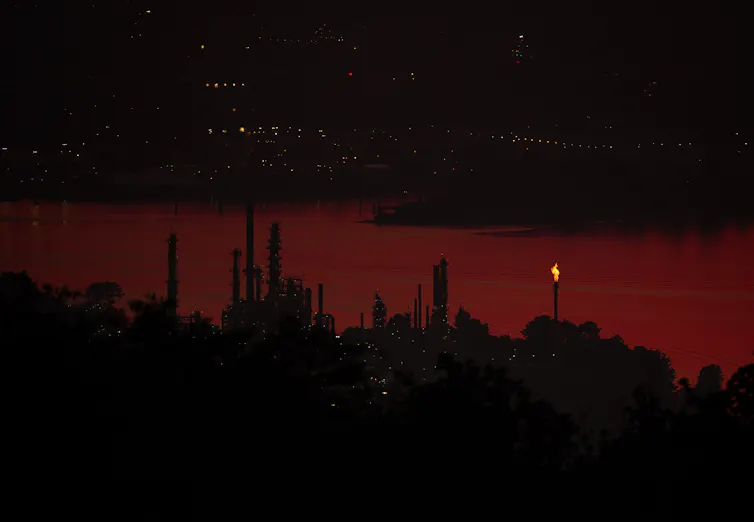 refinería de petróleo con bengalas cerca del río con paisaje urbano de fondo al atardecer.