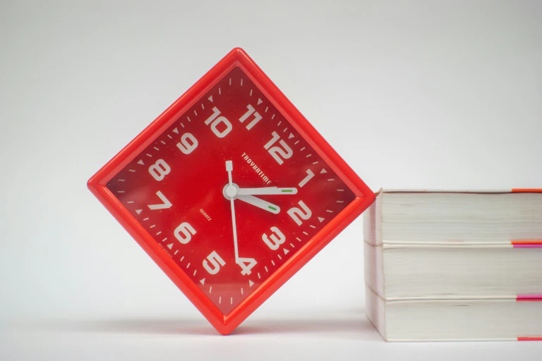 Square red clock leaning against a stack of books