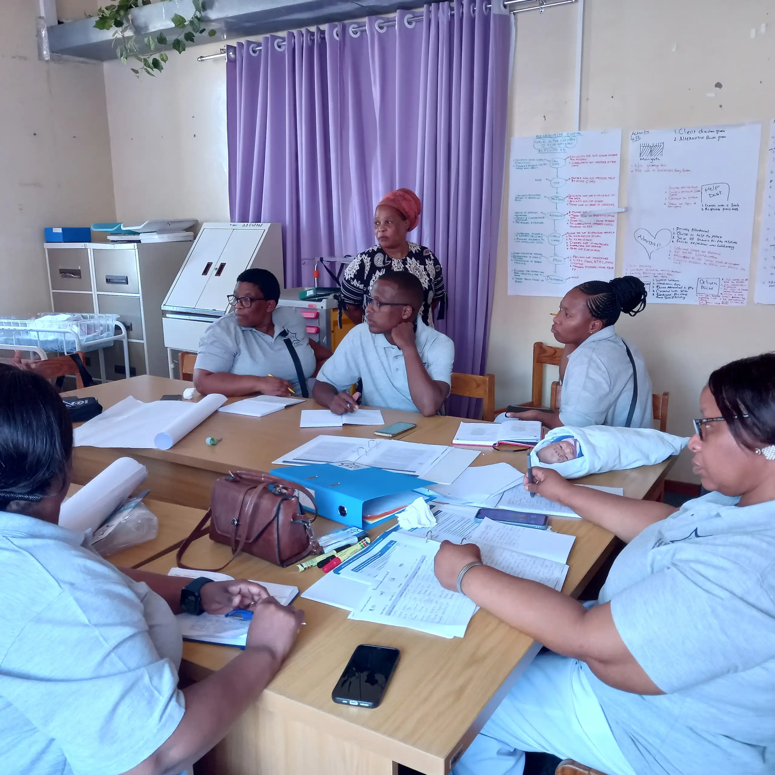 Health workers in a meeting around a table with notebooks; a 'newborn' doll is on the table too