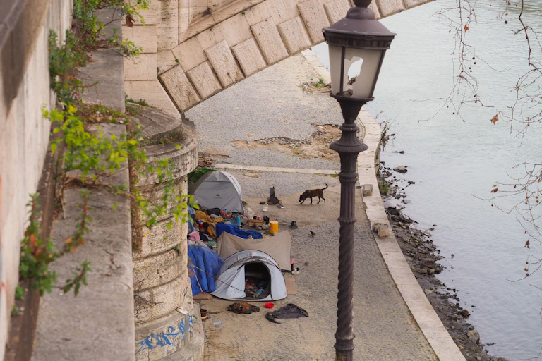 tents of homeless people by bridge next to river, black lamppost in foreground