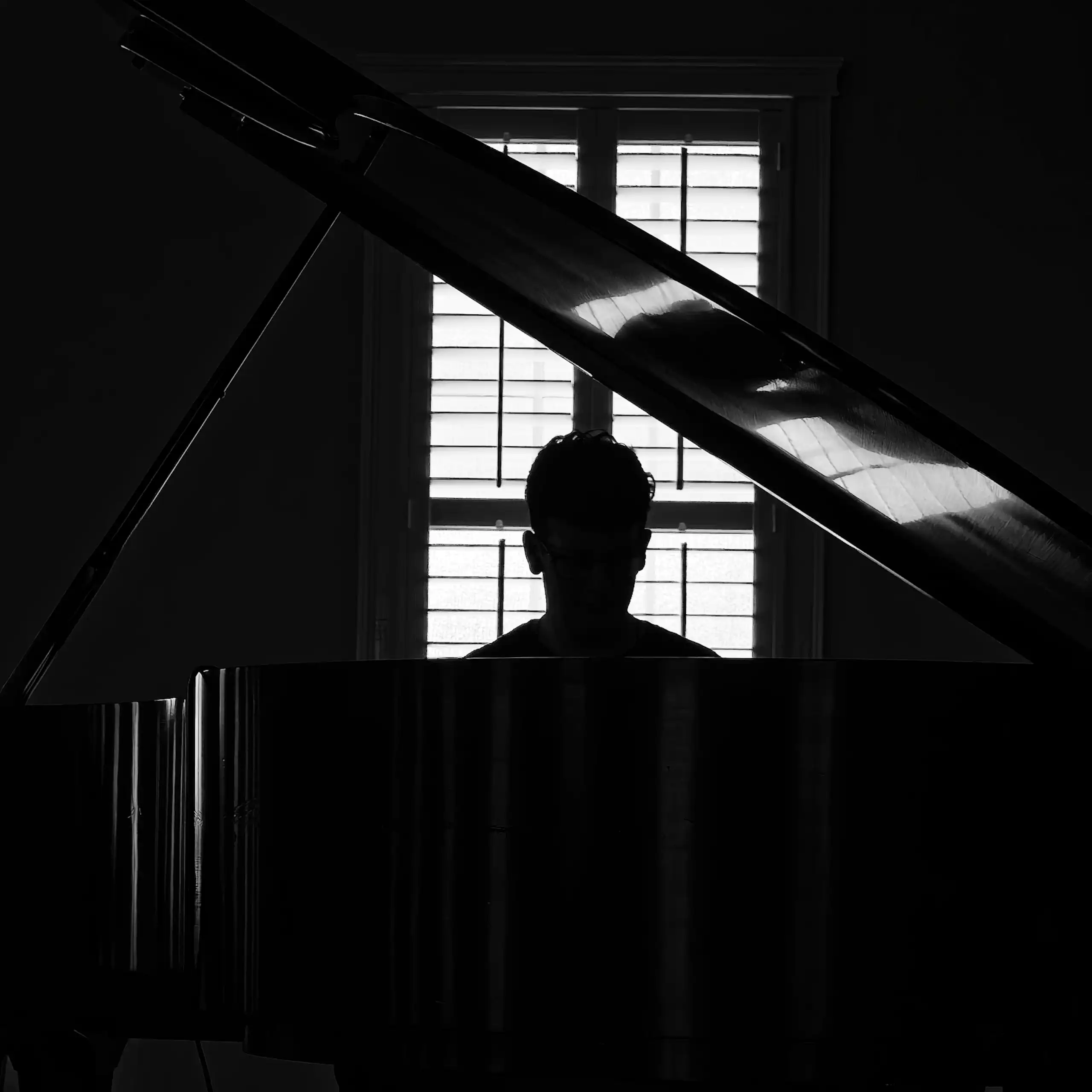 A black and white photo illustration of a young person in silhouette, sitting at a grand piano in a dark room with a window framing them with shutter lines forming bars.