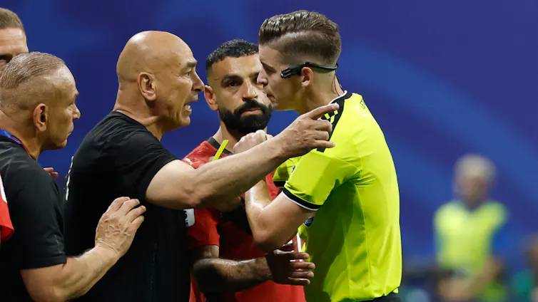 Two men stand face to face with one pointing and speaking as a third man holds his arm out between the first two.