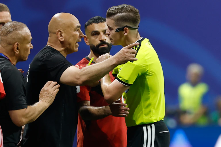 Two men stand face to face with one pointing and speaking as a third man holds his arm out between the first two.