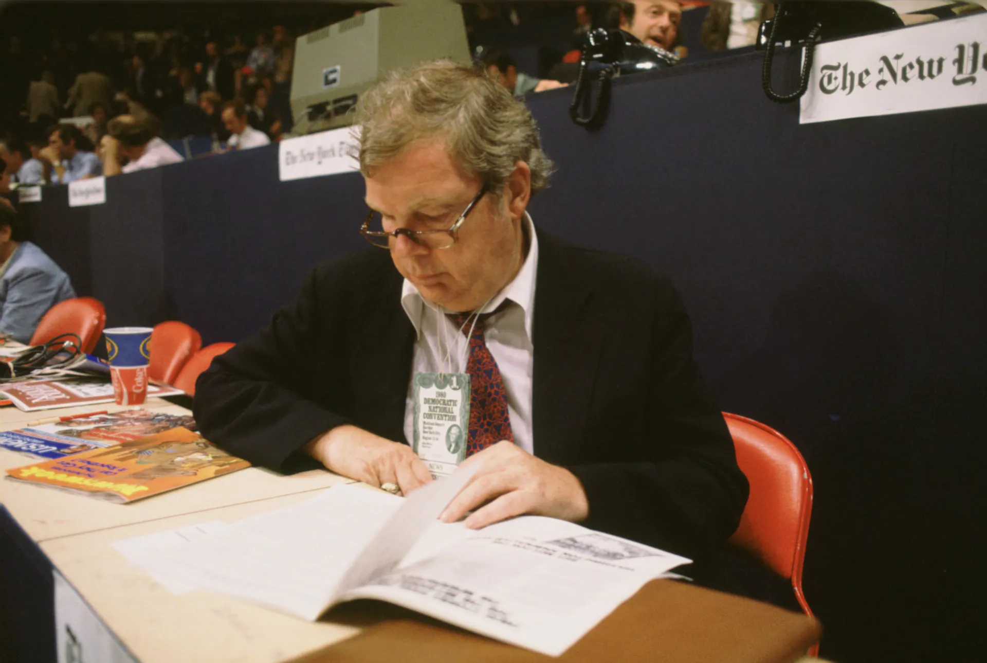 A dark-haired man with glasses sitting at a long table, reading papers.