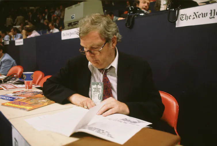 A dark-haired man with glasses sitting at a long table, reading papers.