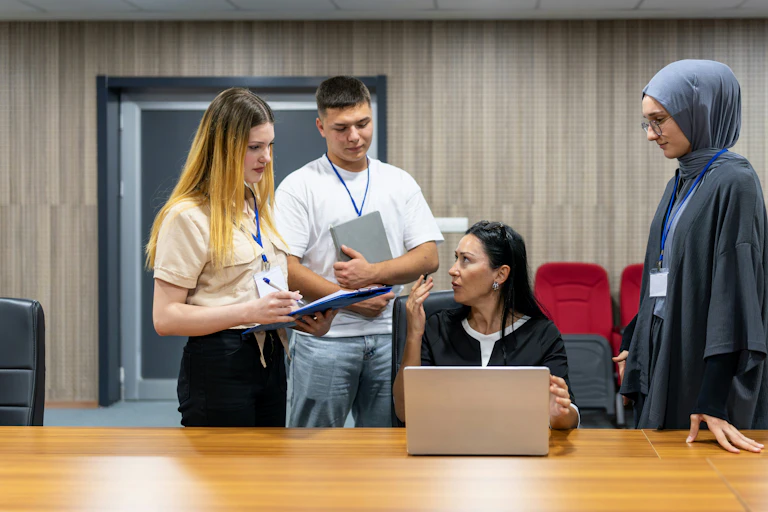 A person with a laptop at a desk flanked by young people with lanyards, one wears a hijab.