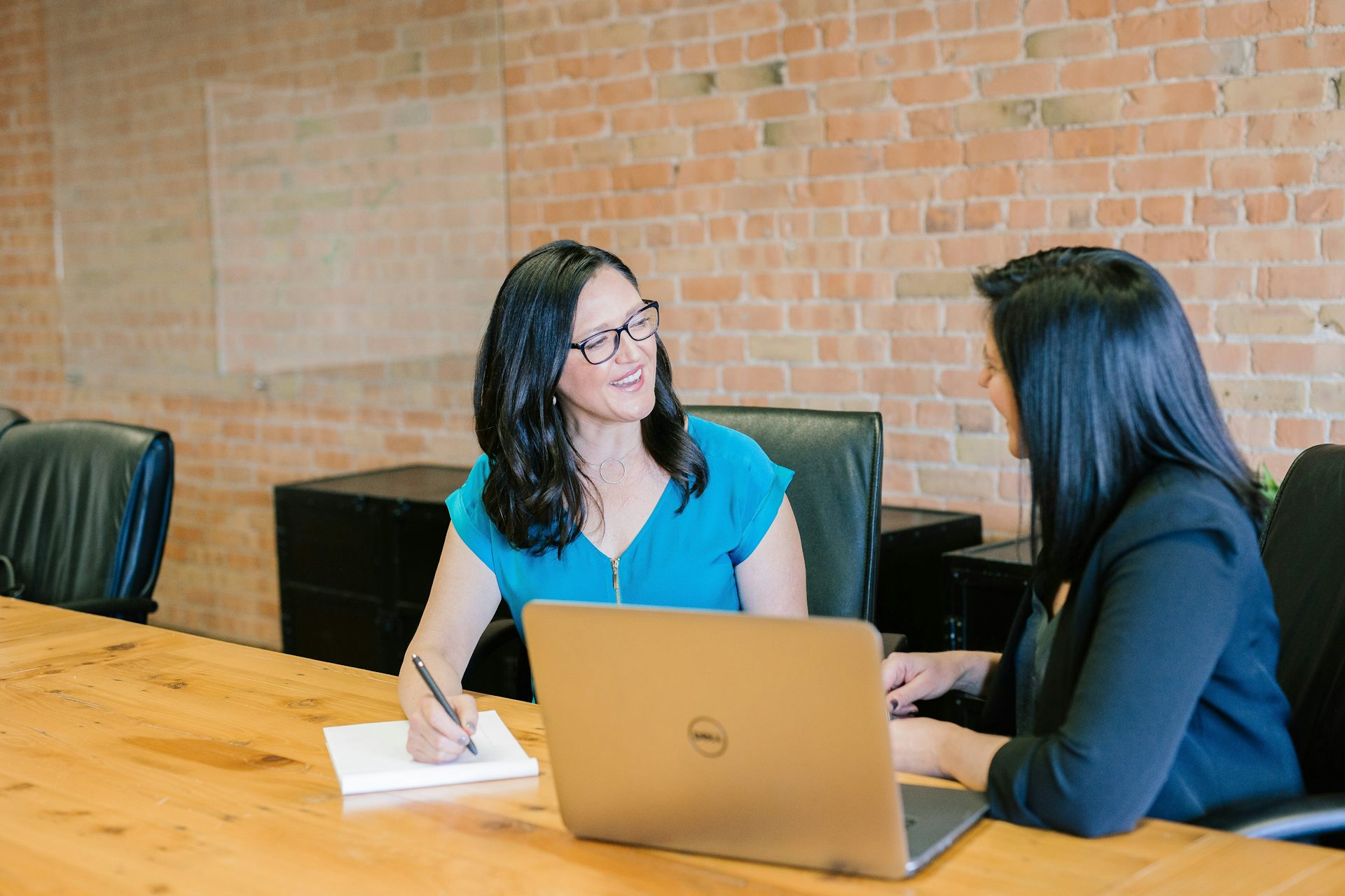 Two women talk with each other at a table with a laptop and notepad next to them.