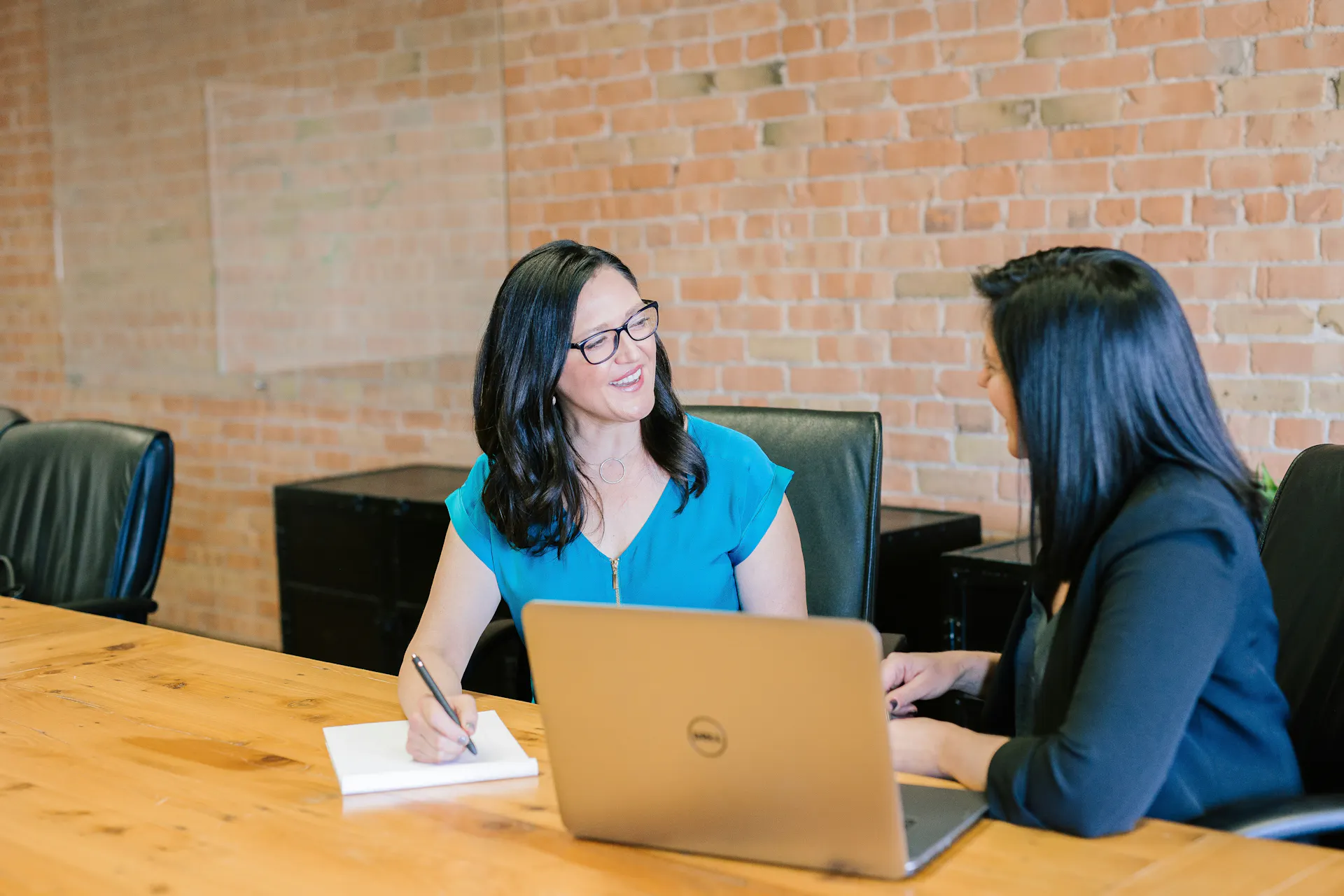 Two women talk with each other at a table with a laptop and notepad next to them.
