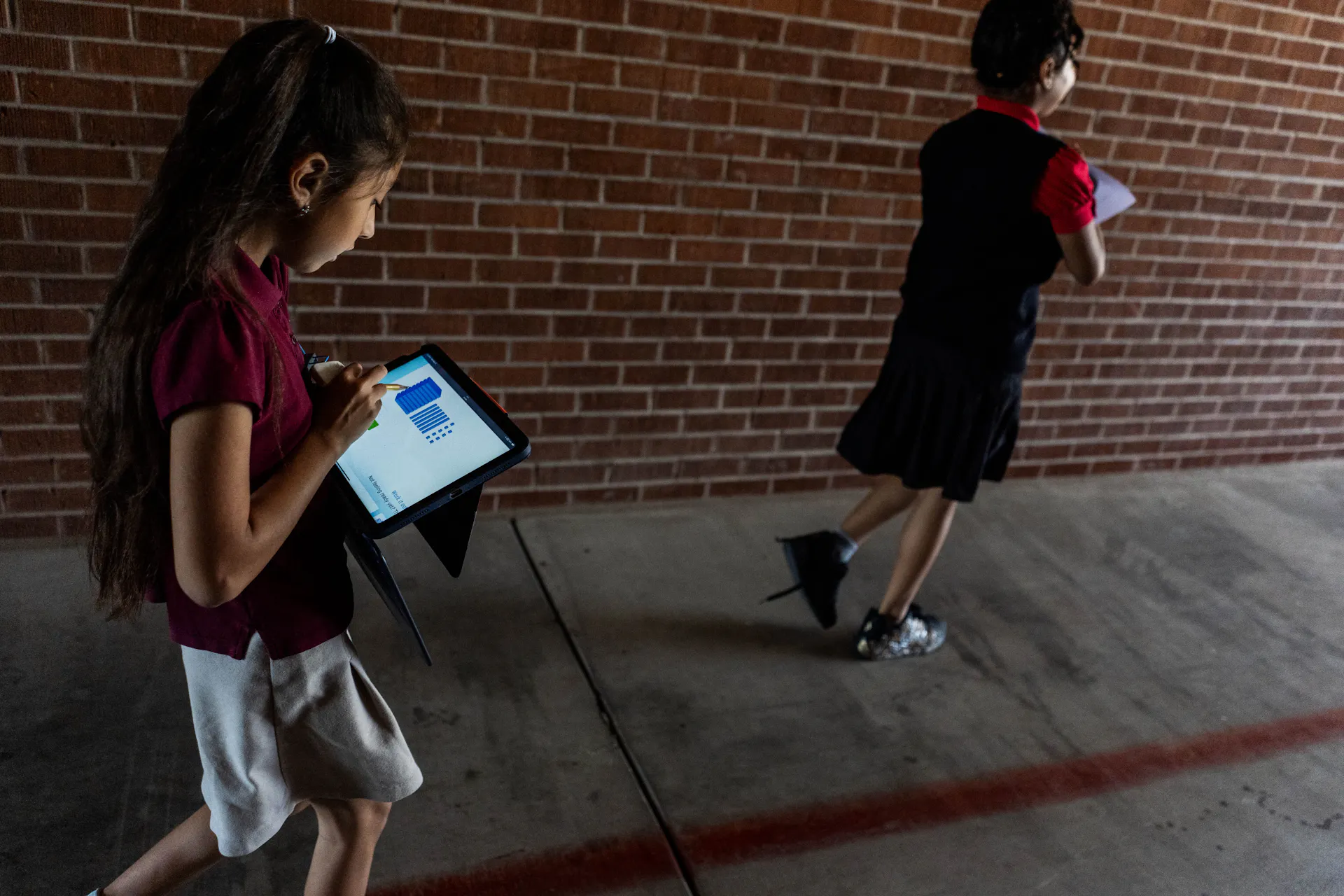 Two children wearing maroon shirts walk near a red brick wall and look down at iPads.