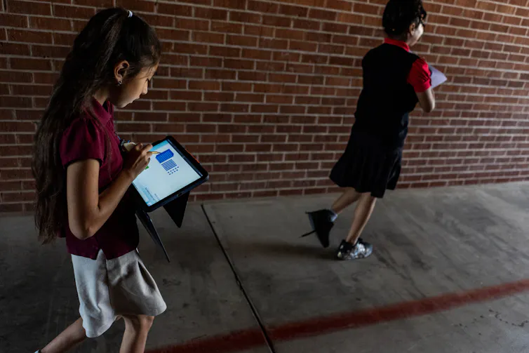 Two children wearing maroon shirts walk near a red brick wall and look down at iPads.
