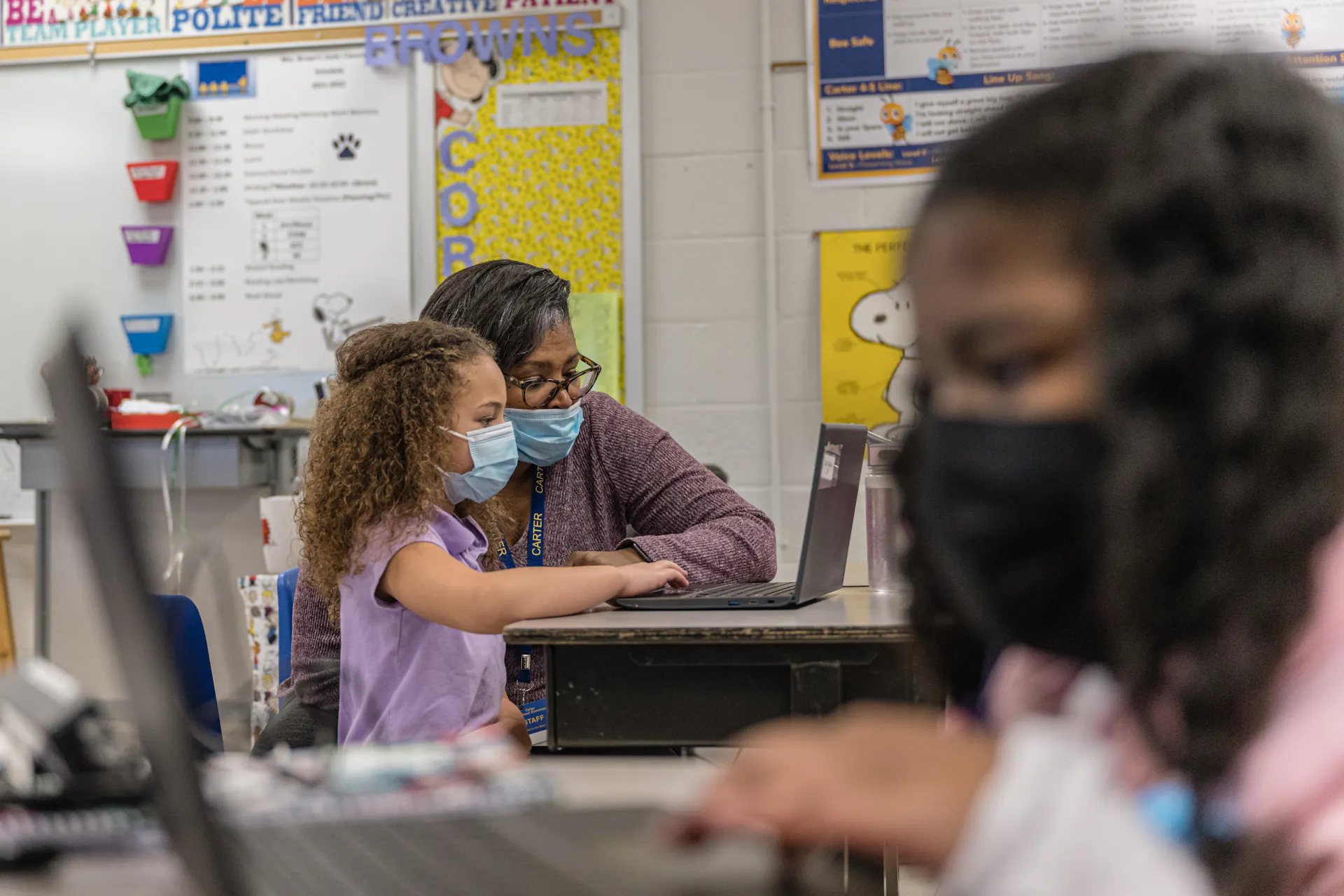 Two girls wearing face masks sit in opposite directions and look at computer screens. One of the girls sits next to an adult, also wearing a mask.