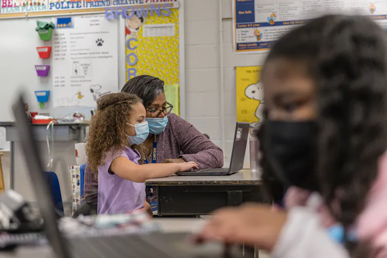 Two girls wearing face masks sit in opposite directions and look at computer screens. One of the girls sits next to an adult, also wearing a mask.