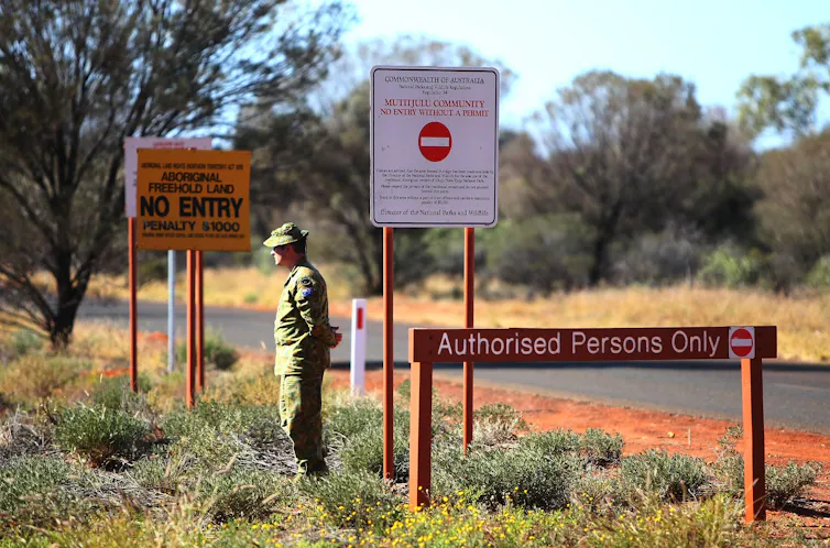 A soldier stands in the Australian outback surrounded by signs saying a permit is needed for entry.