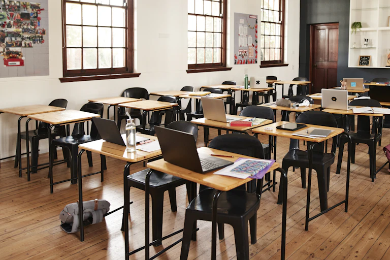An empty classroom has laptops open on a series of desk lined up in rows.