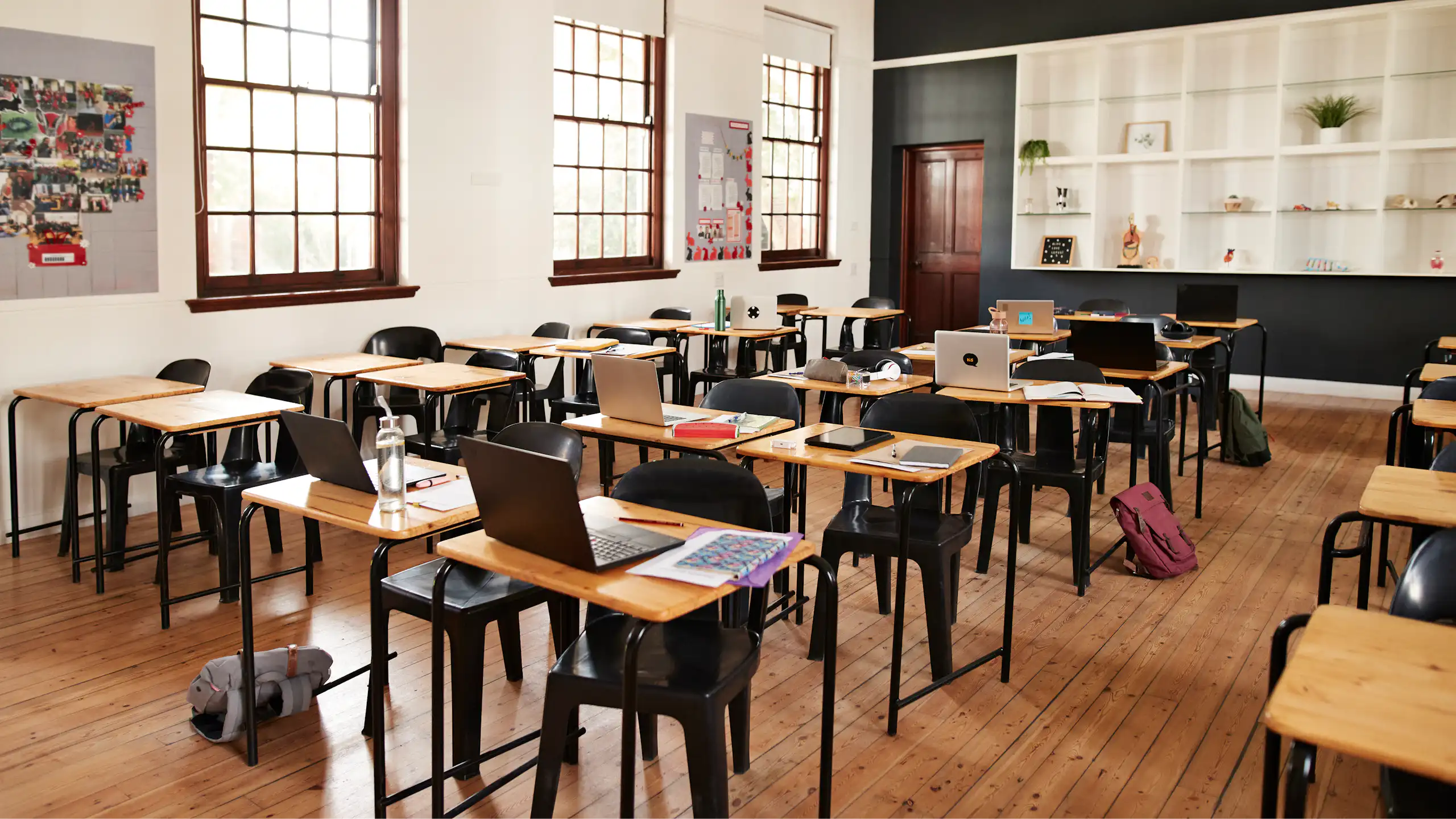An empty classroom has laptops open on a series of desk lined up in rows.