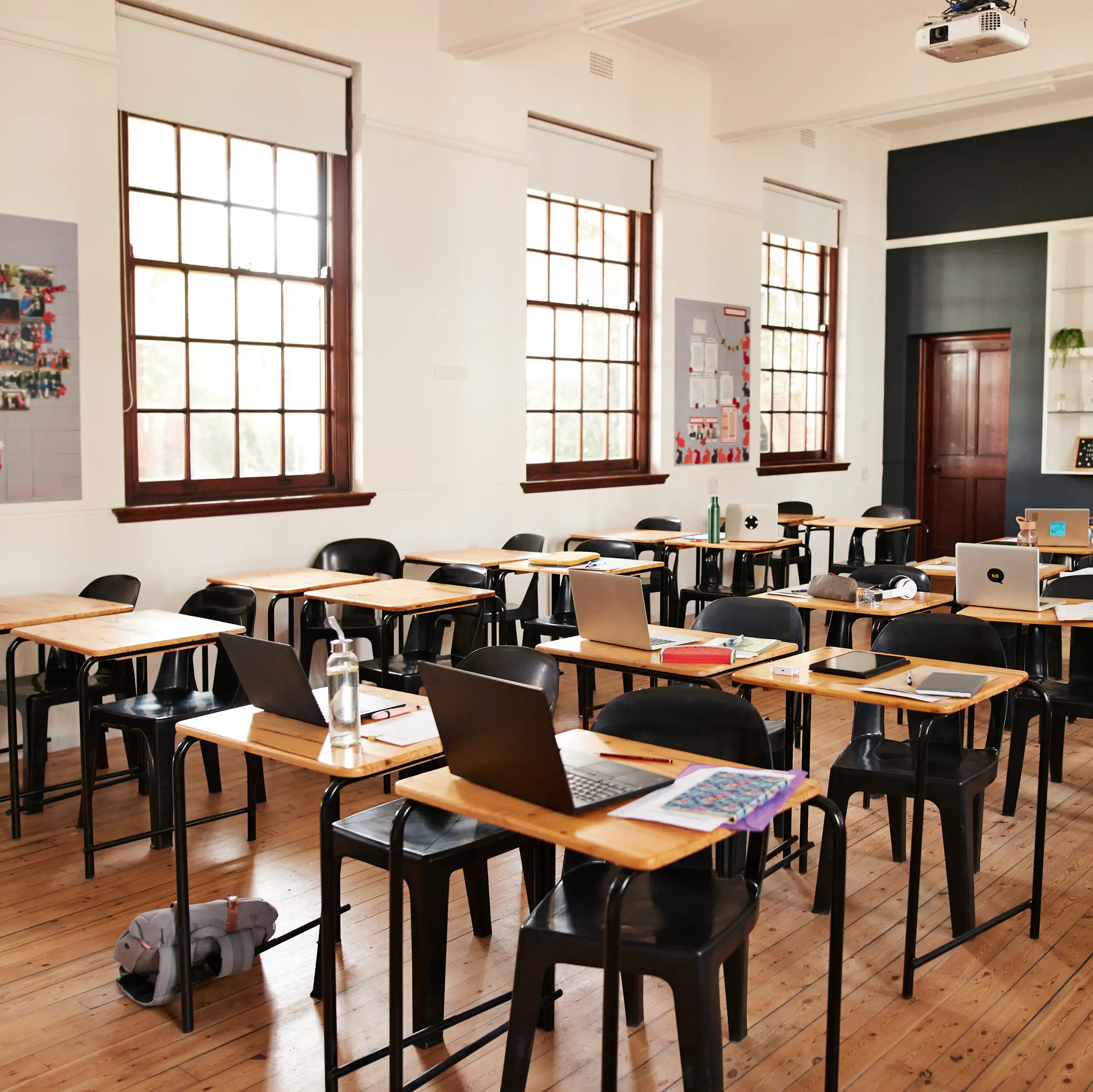 An empty classroom has laptops open on a series of desk lined up in rows.