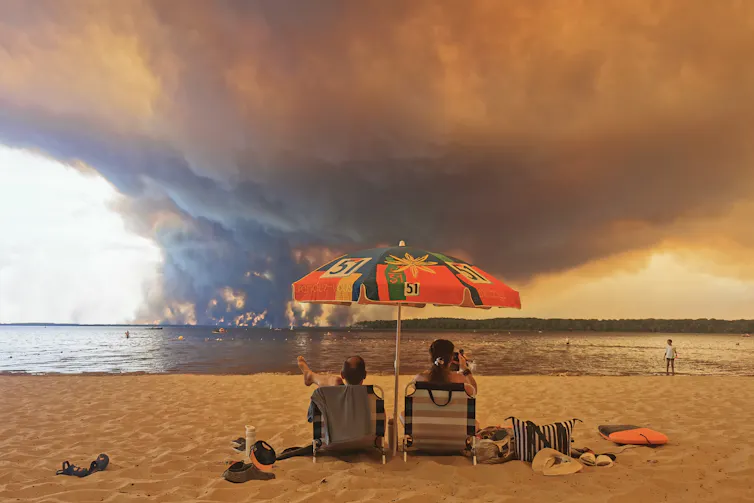 two people sit at lakeside beach watching plume of wildfire smoke in distance.