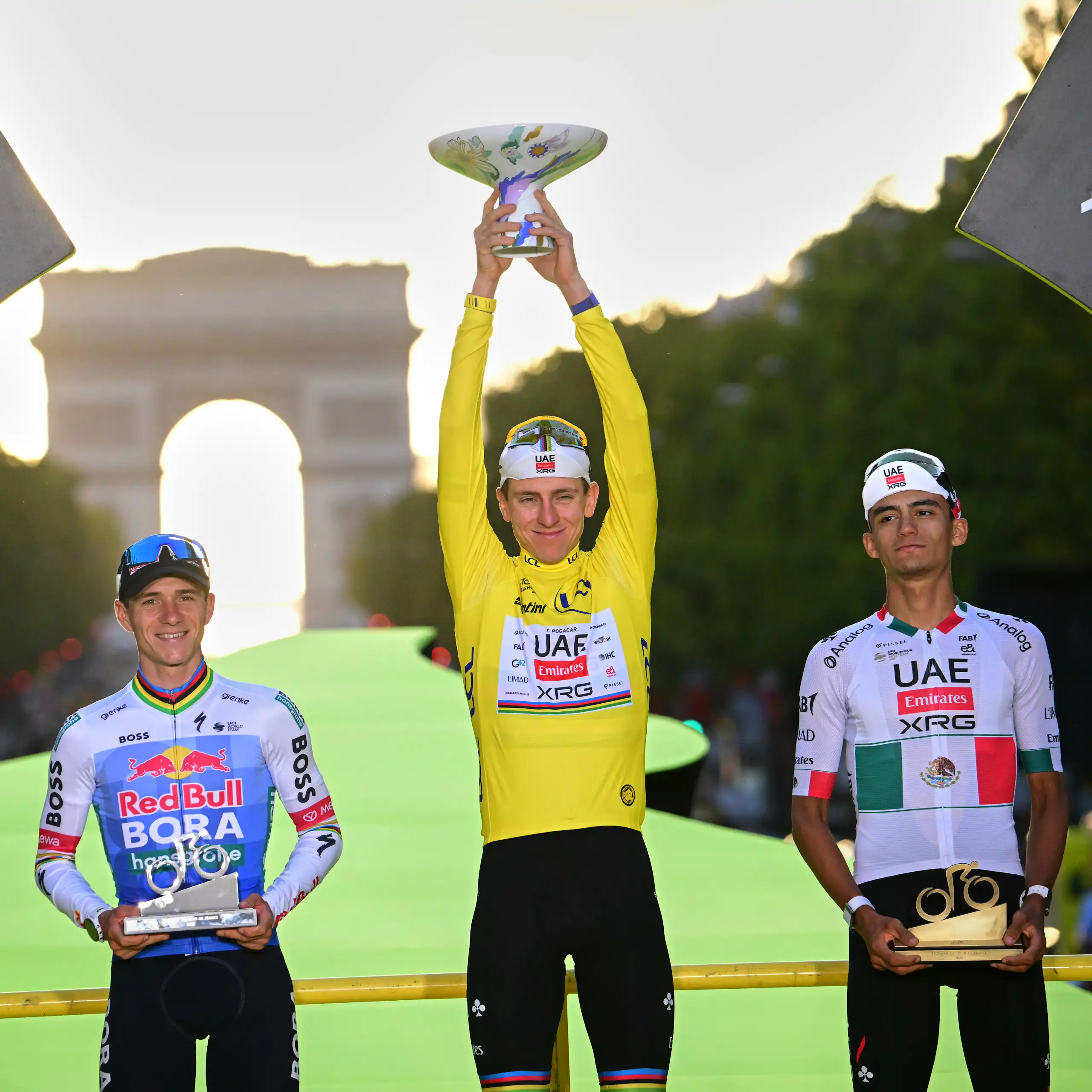 Three men stand on a podium, one holding up a trophy