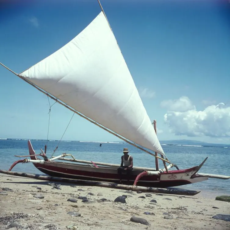 A fisher sits on the edge of a Balienese fishing boat on a beach.
