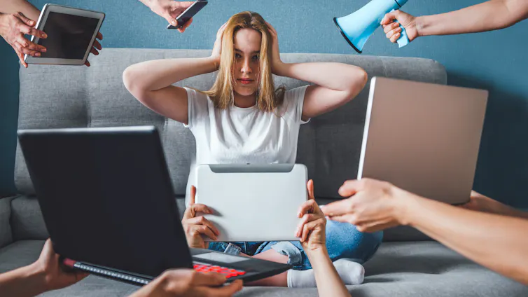 Woman sit on sofa and puts hands over her ears while lots of digital devices and a loudspeaker are held around her