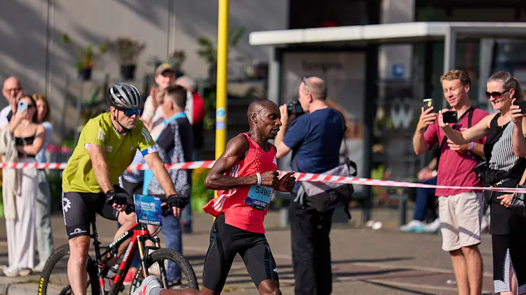 An African man runs on the road followed by a man on a bicycle as people behind a taped barrier take photos of the runner on their phones.