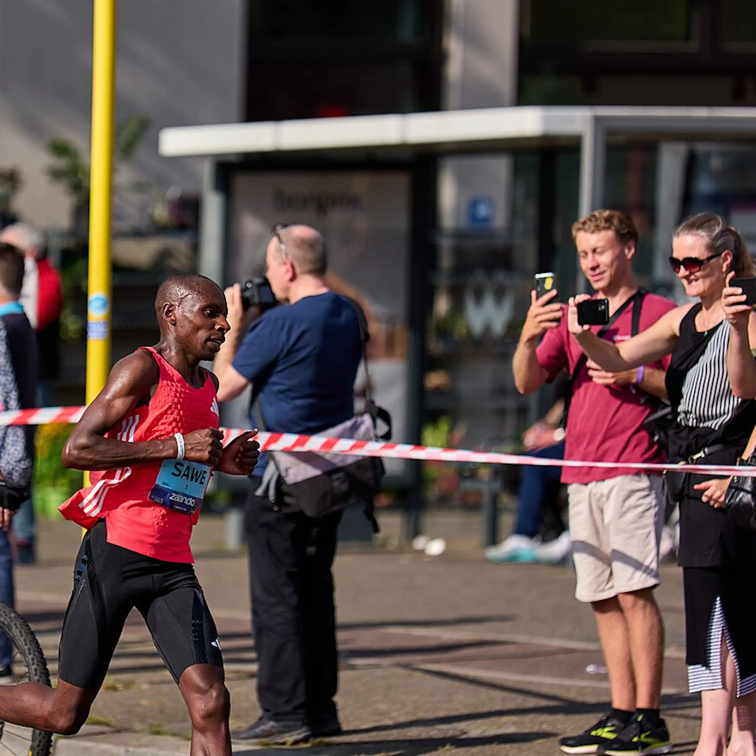 An African man runs on the road followed by a man on a bicycle as people behind a taped barrier take photos of the runner on their phones.