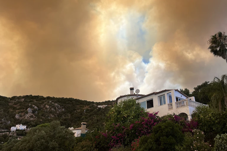 A house on a mountainside, with vegetation and smoke from fires in the sky.