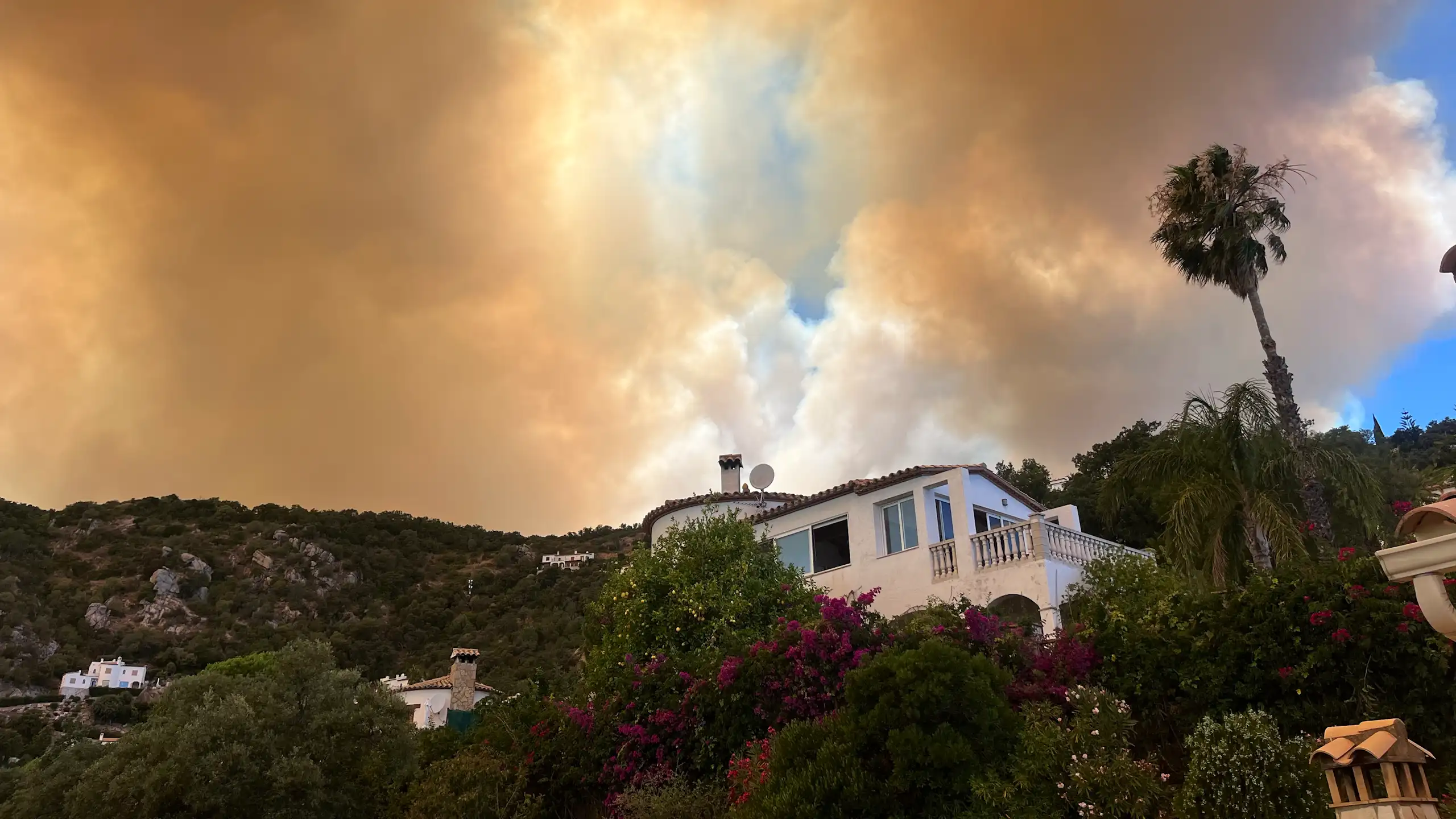 A house on a mountainside, with vegetation and smoke from fires in the sky.