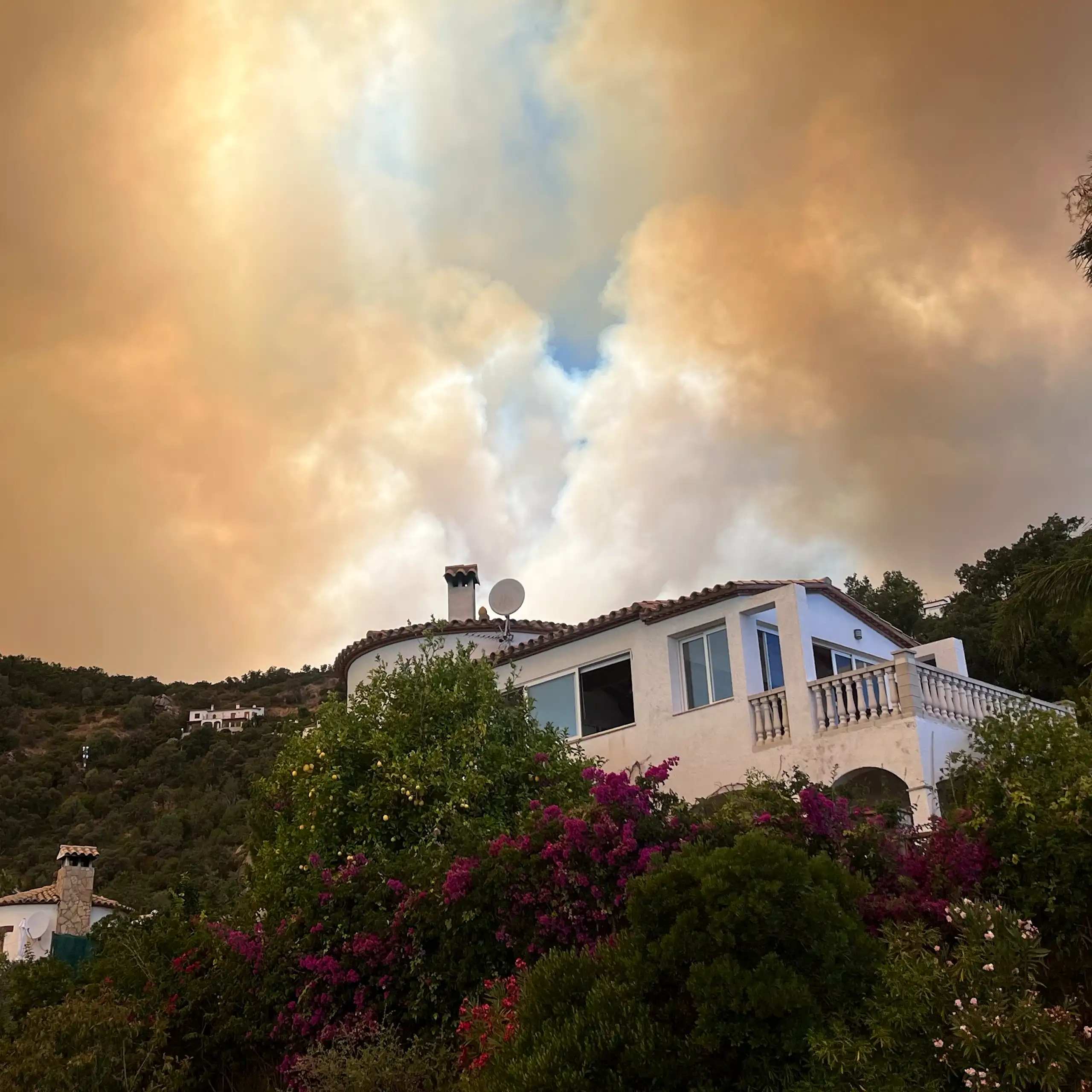 A house on a mountainside, with vegetation and smoke from fires in the sky.